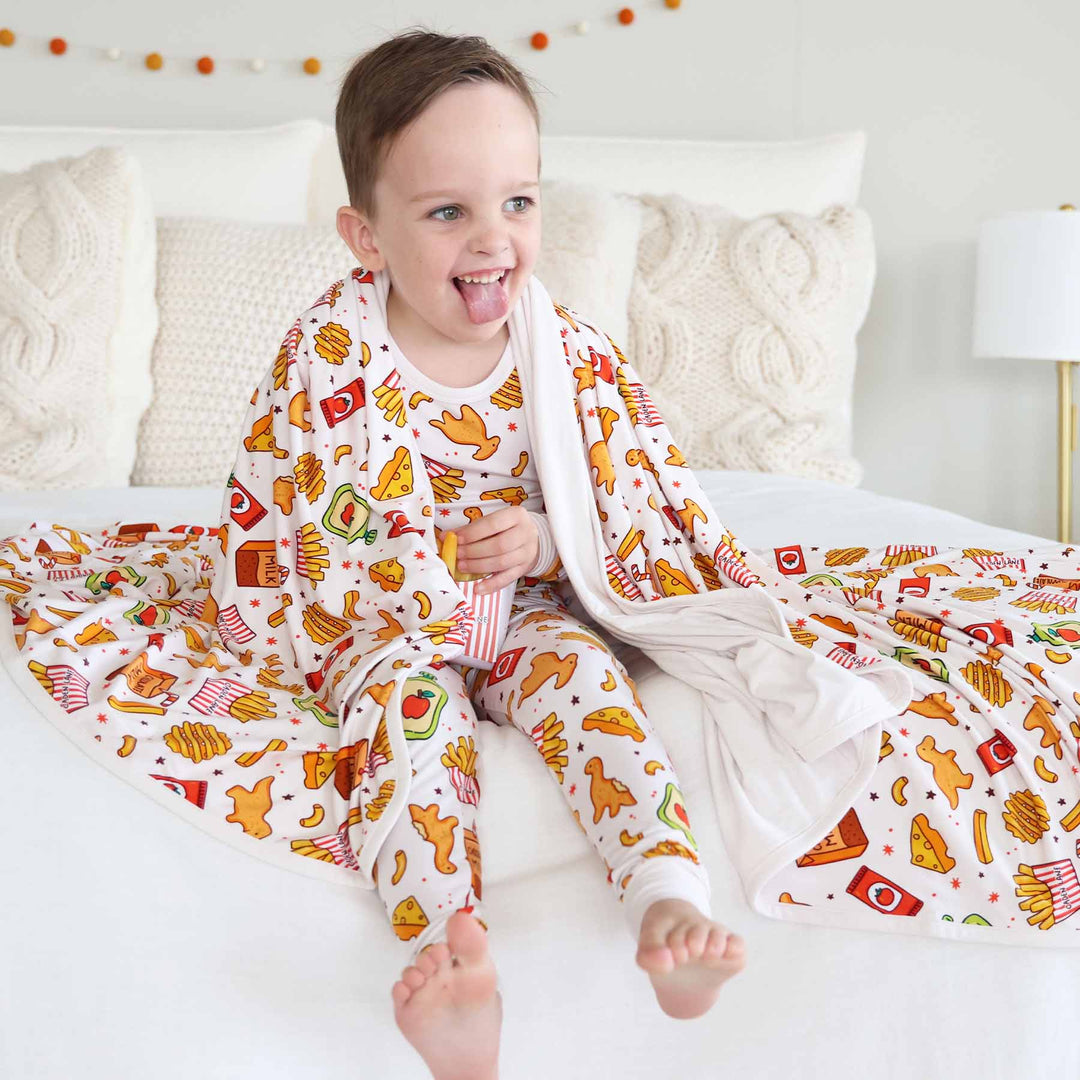 boy sitting on bed with food themed pajamas with a matching bamboo blanket draped over his shoulders 