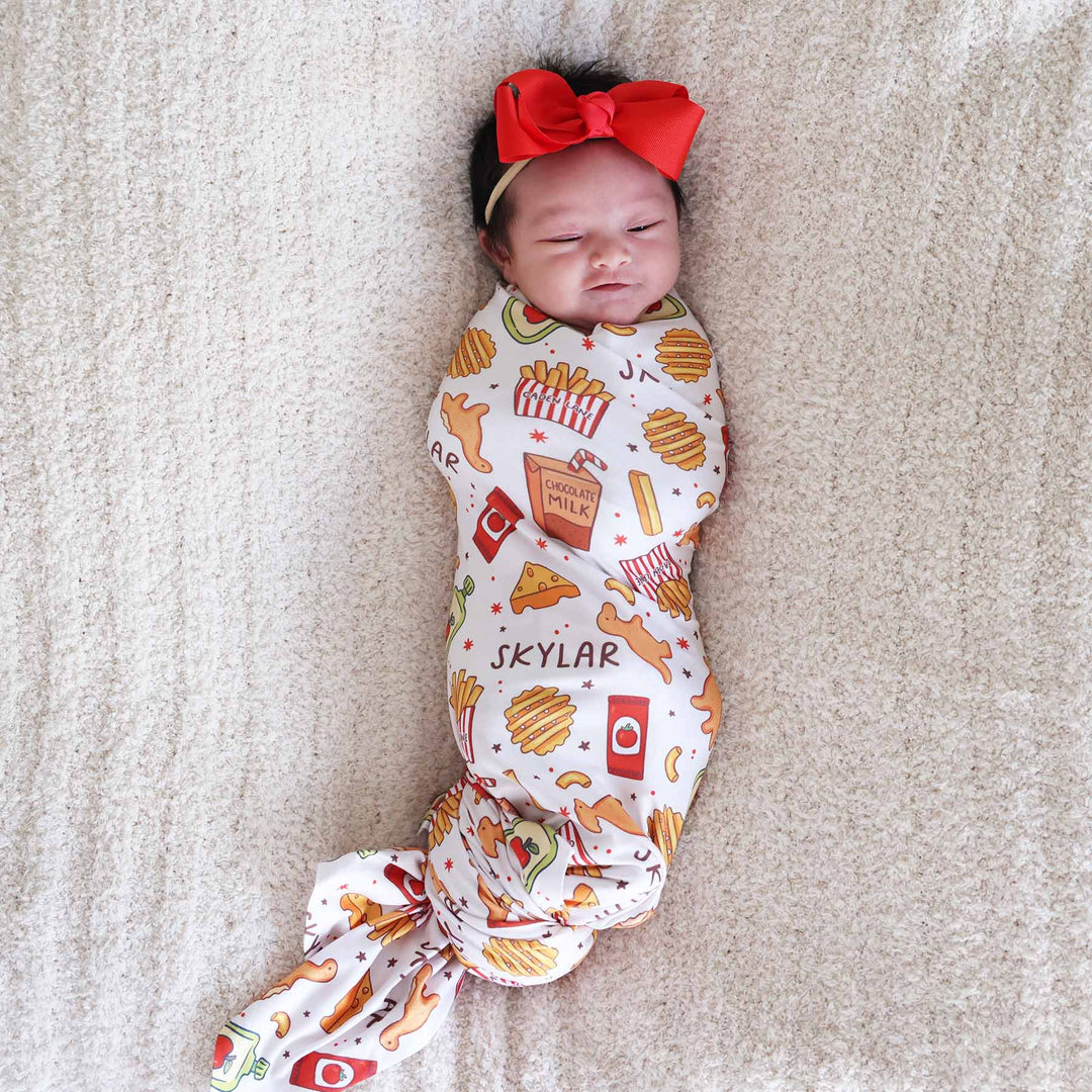 newborn girl wrapped in a food themed personalized swaddle blanket with a matching red bow headband on her head 