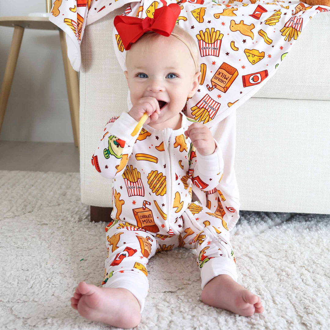 girl sitting on ground in snack themed zip romper pajama with a french fry in her mouth and red ribbon bow headband on her head 