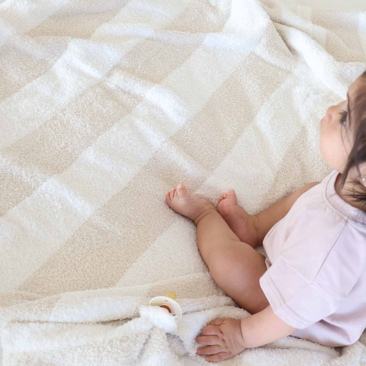 baby girl sitting on a beige and white stripe microfiber blanket next to a matching lovey