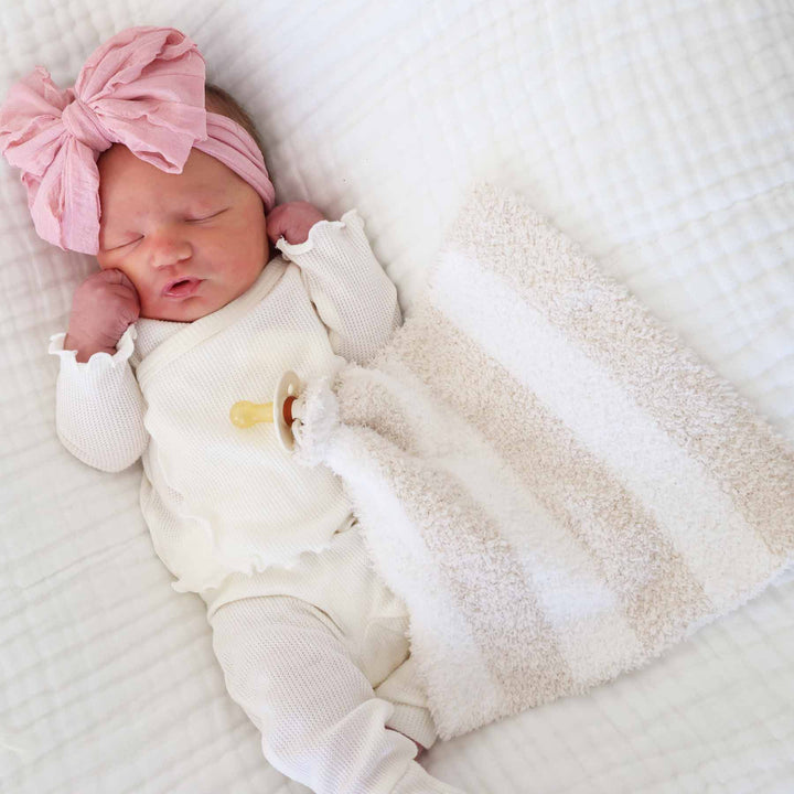 baby laying on top of a white swaddle in a bamboo waffle two piece outfit with a fluffy pink bow with a pacifier attached to a toffee stripe plush lovey on top of her 