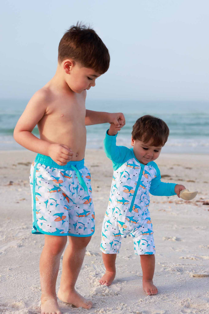 older brother holding little brothers hand on the beach wearing matching shark swimsuits 