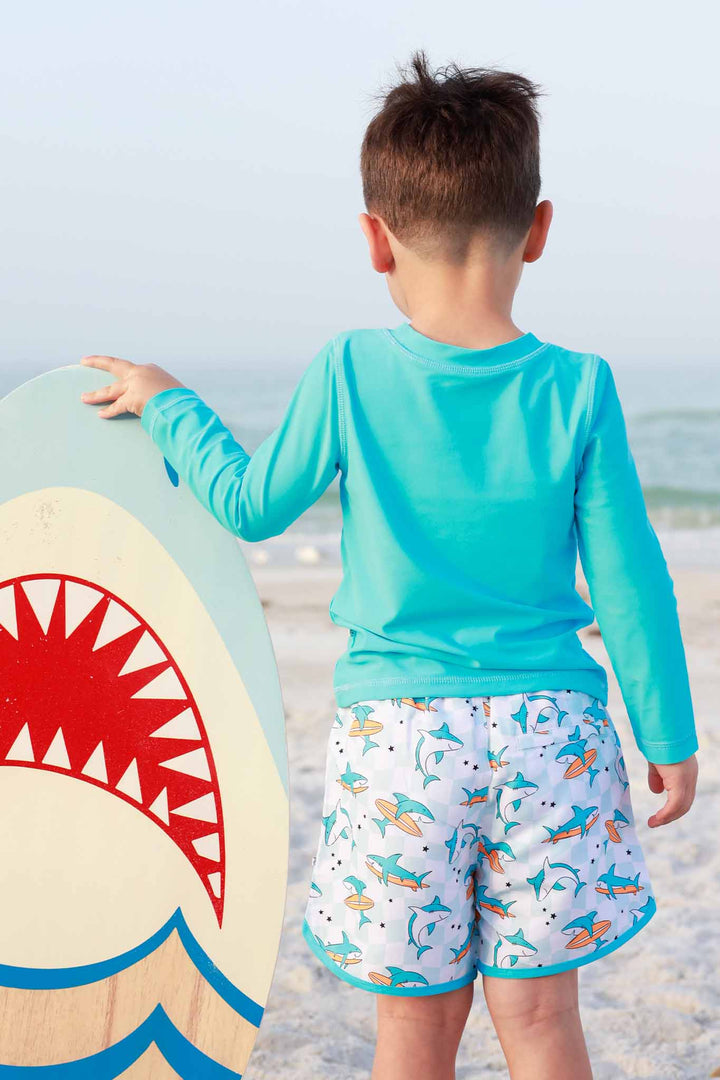 boy standing on beach in surfing shark swim trunks with a long sleeve turquoise rash guard shirt 