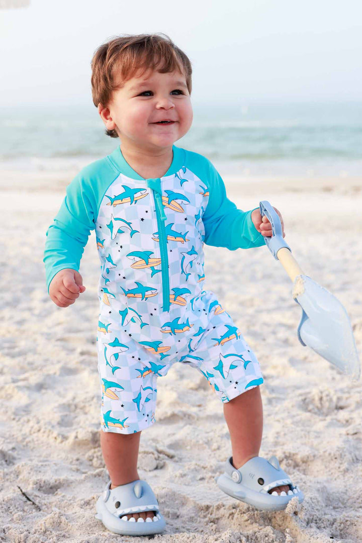 boy holding shovel walking on beach in surfing shark romper shortie swimsuit 