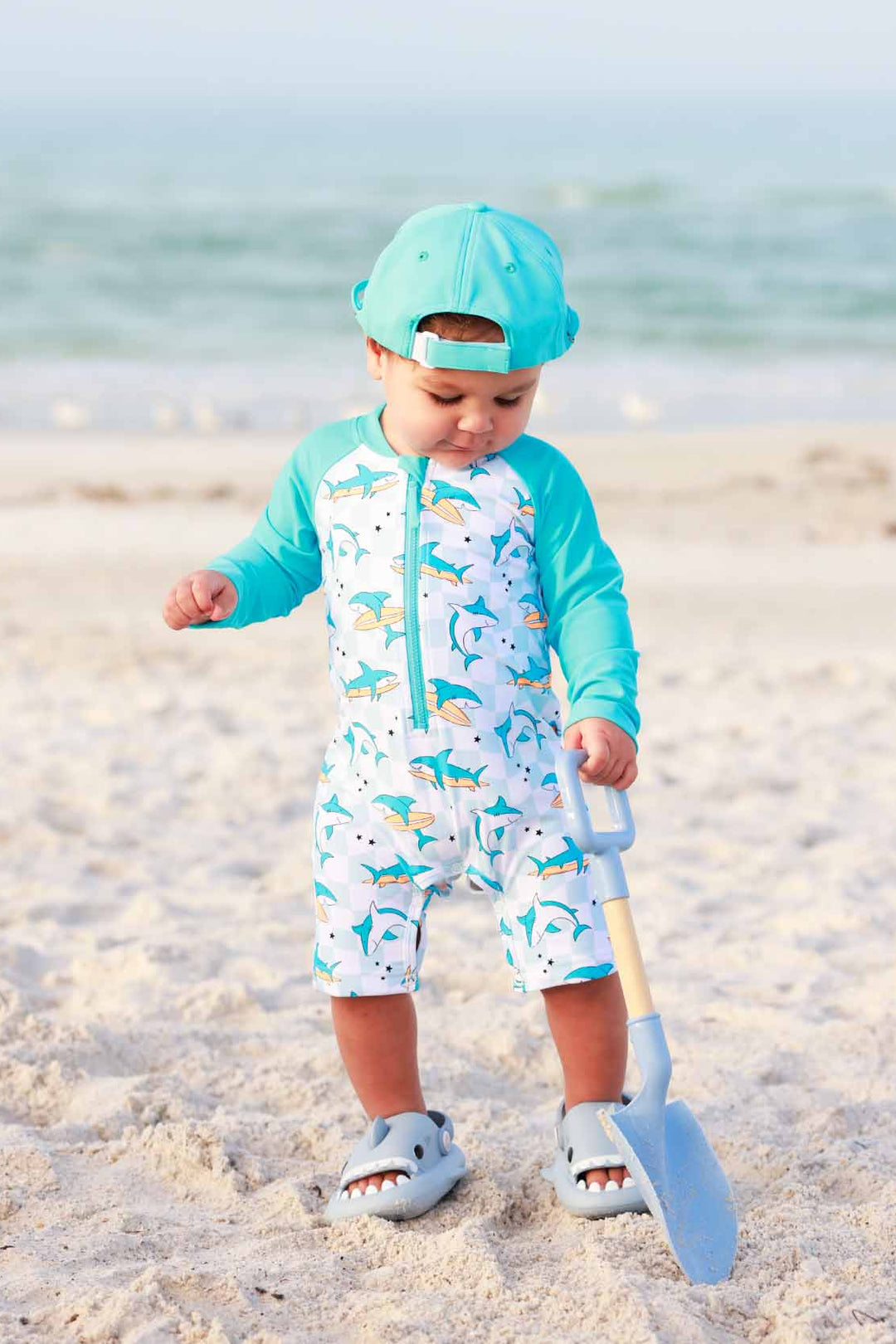 boy playing with beach shovel in surfing shark shortie romper with a matching hat and shark shoes 