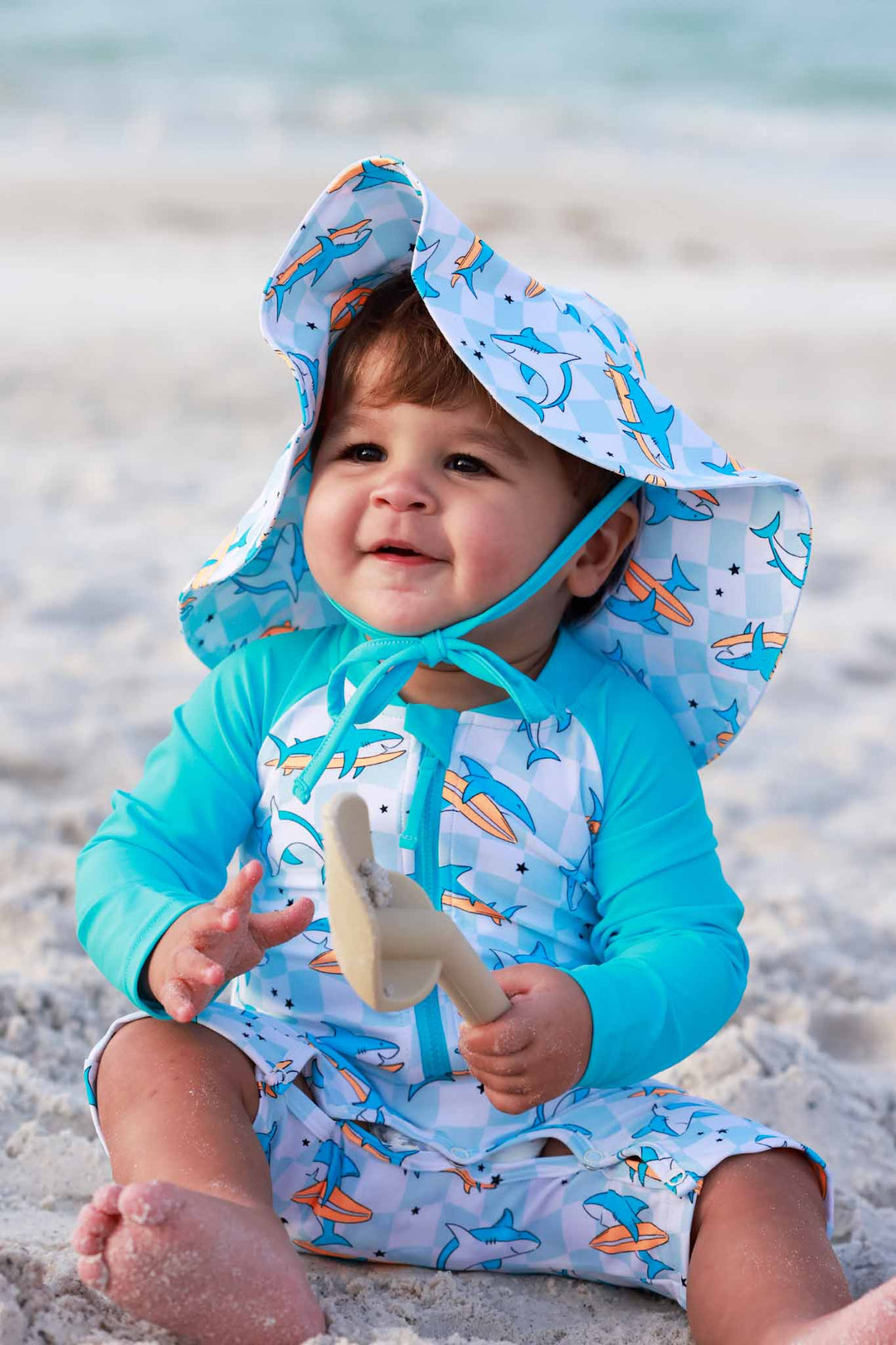 little boy sitting in sand in long sleeve rash guard swim romper shortie in surfing shark print with a matching sun hat 