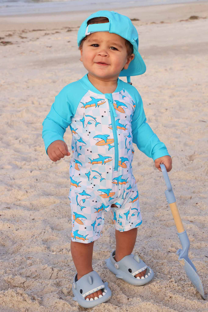 boy standing in sand in surfing shark romper with a backwards matching turquoise surf hat 