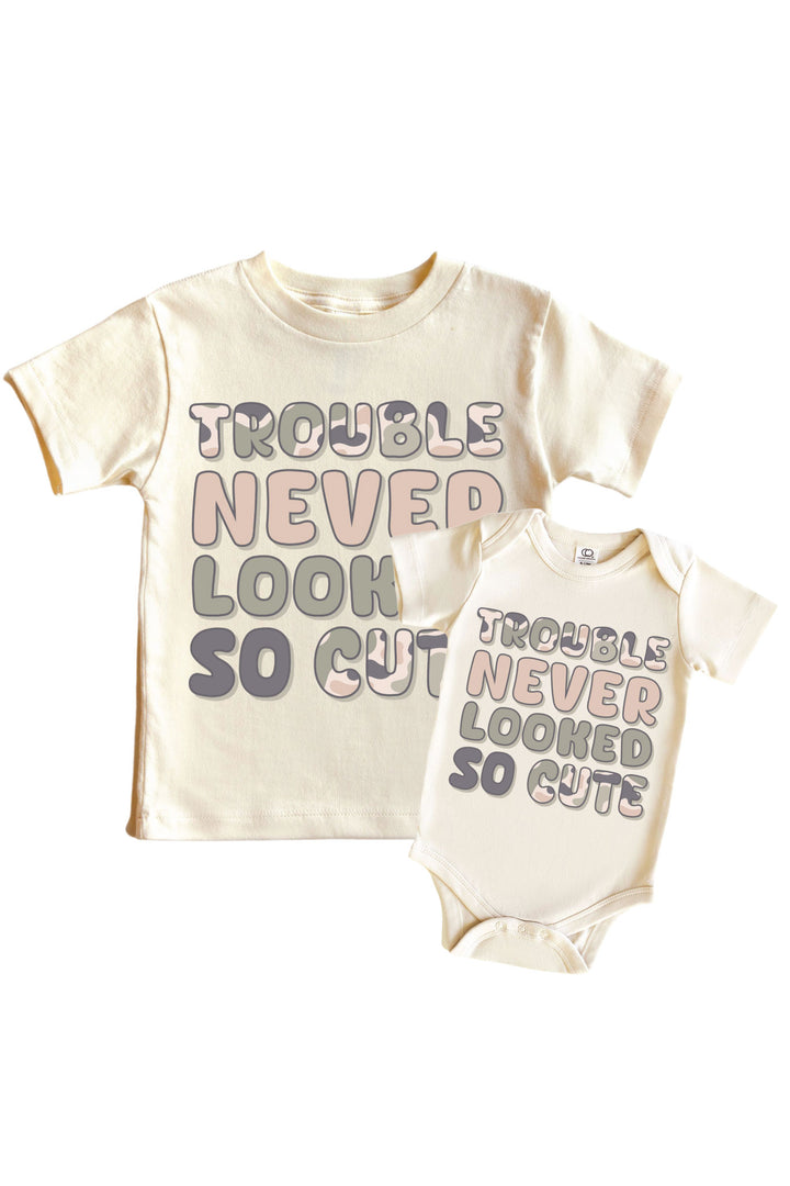 Natural cream kids graphic tee and baby bodysuit with "Trouble Never Looked So Cute" slogan in pink and gray block lettering