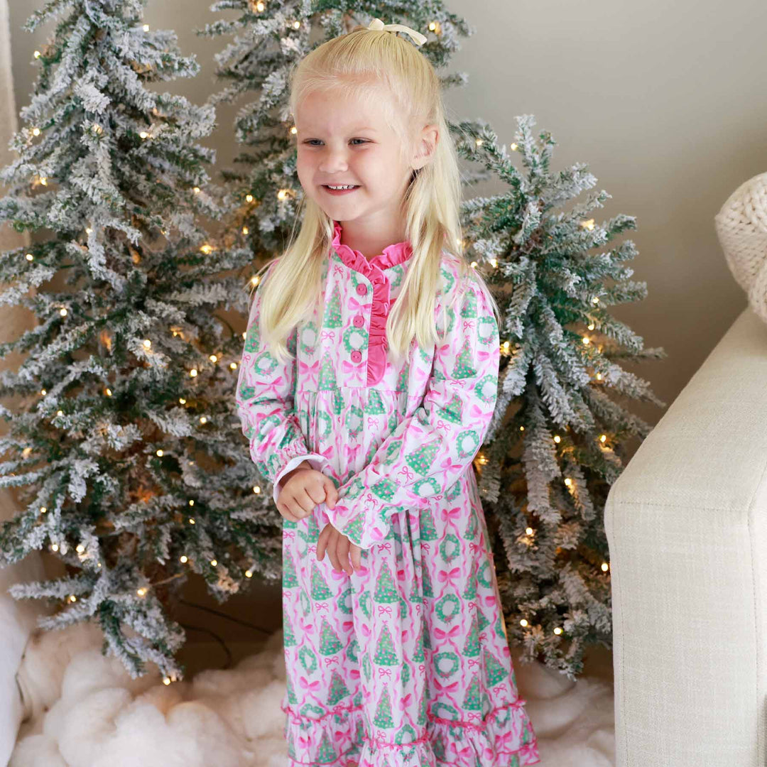 girl wearing long sleeve christmas nightgown with bows, wreaths and christmas trees standing in front of a christmas tree