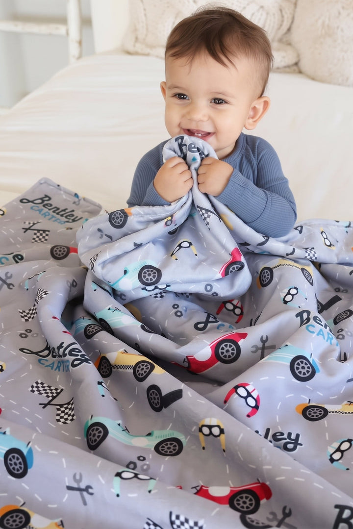 A baby sitting on a white bed, holding a Caden Lane light grey blanket printed with colorful toy cars and the name 'Rebel' in script.