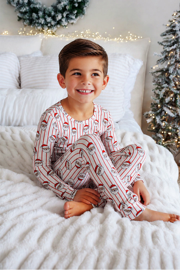 A young boy smiling while sitting on a white bed, wearing a long-sleeve holiday-themed pajama set with red and white stripes and Santa Claus illustrations. The background features a cozy bedroom setting with a decorated Christmas tree and warm string lights.