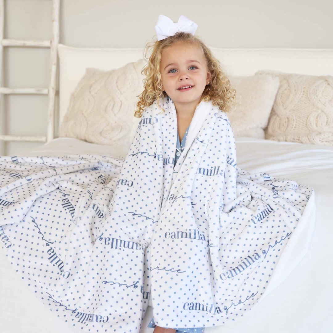girl sitting on bed wrapped in a personalized blanket that is white with blue polka dots 