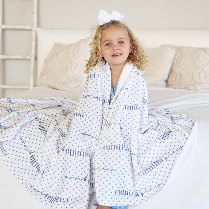 girl sitting on bed wrapped in a personalized blanket that is white with blue polka dots 