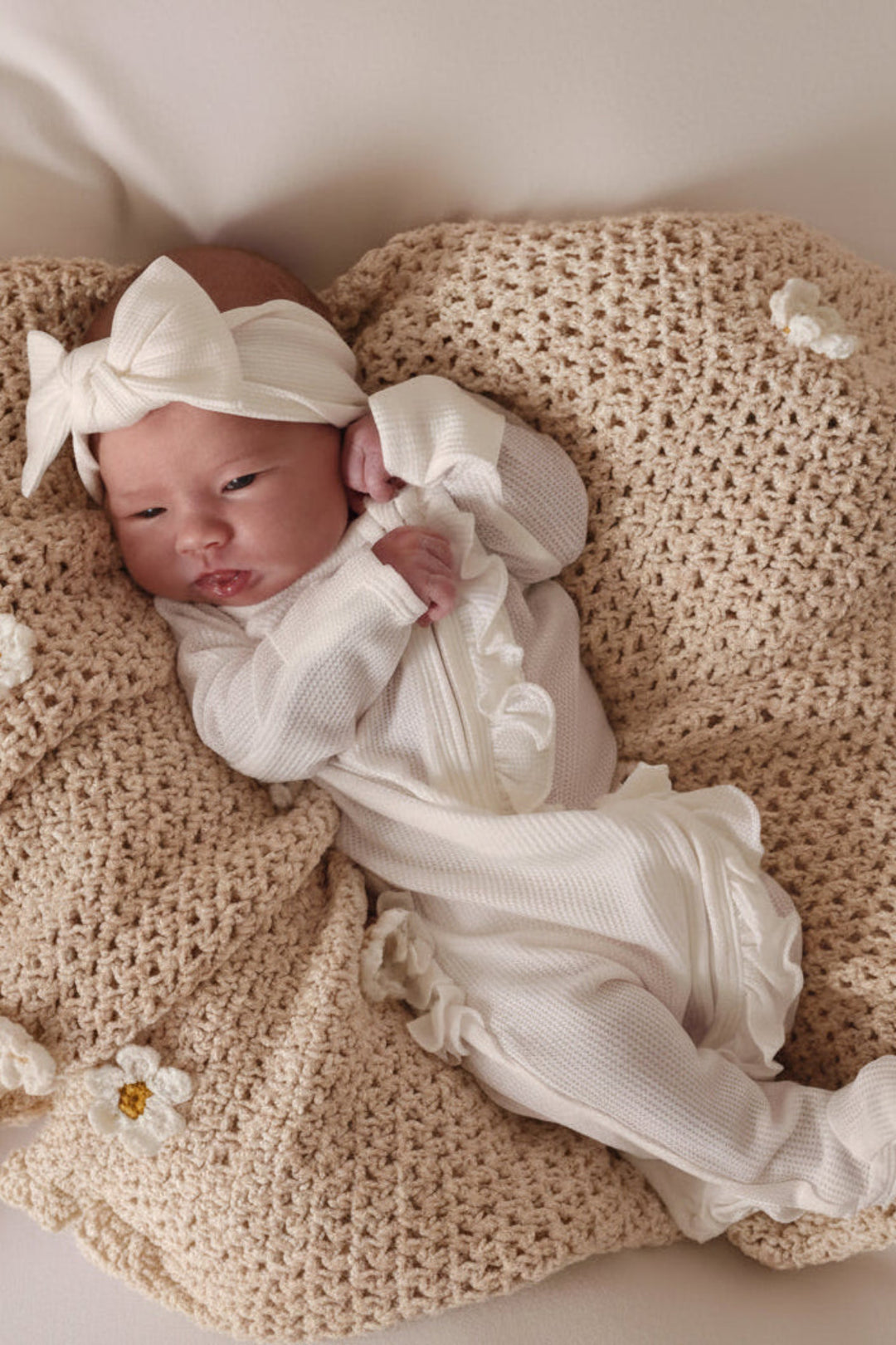 Newborn baby in white outfit and headband lying on a textured beige blanket.